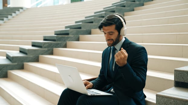 Confident smart business man celebrate successful business idea while listen music. Happy manager project working by using laptop while wearing headphone and suit. Investor sitting at stair. Exultant.