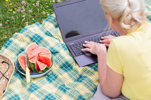 young blonde woman with a laptop lies on a blanket in the green grass on a sunny day, watermelon summer picnic, remote work, virtual chat, High quality photo
