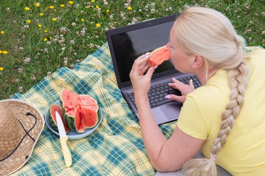 young blonde woman with a laptop lies on a blanket in the green grass on a sunny day, watermelon summer picnic, remote work, virtual chat, High quality photo