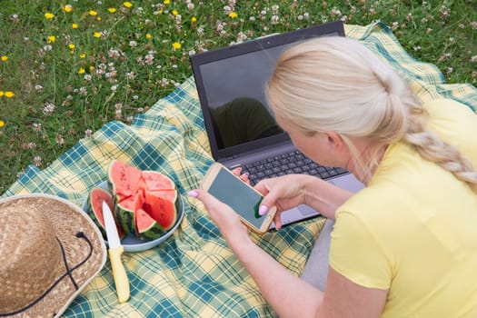 young blonde woman with mobile phone and laptop lies on a blanket in green grass on a sunny day, watermelon summer picnic, remote work, virtual chat, green screen, app mockup, high quality photo