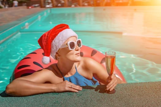 Woman pool Santa hat. A happy woman in a blue bikini, a red and white Santa hat and sunglasses poses near the pool with a glass of champagne standing nearby. Christmas holidays concept