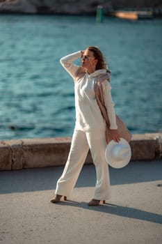 Happy blonde woman in a white suit and hat posing at the camera against the backdrop of the sea.