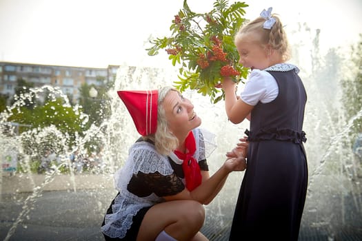 Young and adult schoolgirl on September with flowers having fun near water of fontain. Generations of schoolchildren, pioneer of USSR and October girl in modern uniform of Russia. Mom and daughter