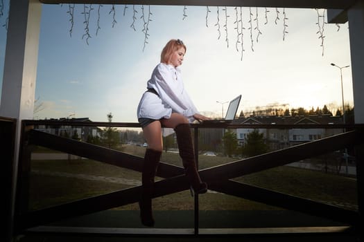 Beautiful redhead moden girl working by laptop in gazebo on autumn, summer or spring day. Businesswoman, student, freelancer or manager works outdoors in natural landscape in a village, city or town
