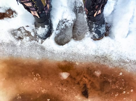 Feet of Hunter or fisherman in big warm boots on winter day on snow. Top view. Fisherman on the ice of a river, lake, reservoir on spring day with melting ice. Dangerous fishing in autumn