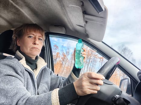 Portrait of female driver in solo journey. Adult mature woman holding steering wheel and bottle of water. Drinking while driving for to stay awake. Lady girl drinks alcohol vodka or gin while driving