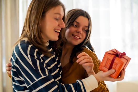 Two happy young girls laugh, friends hug and give each other gifts during the holiday. Women surprise and congratulate on the anniversary of their relationship in their cozy home.