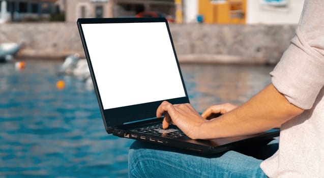 A girl types on a laptop keyboard, sits and works while traveling against the background of a seascape, a bay with the sea on a sunny day, mockup with a white screen, close up view on female hands.