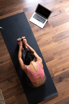 Top view of young woman practicing yoga forward fold on yoga mat with laptop. Vertical image. Fitness and technology concept