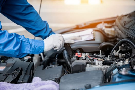 Skilled Asian mechanic in blue workwear jotting down notes on a paper while repairing a car in an auto repair shop. Close-up shot of hand.