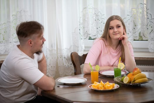 A man and a woman sit at a table in the kitchen. In the background you can see a window and a curtain. There are dishes, drinks and fruit on the table. The woman is wearing a pink sweatshirt.