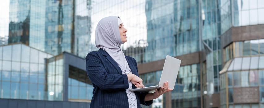 Thoughtful business woman in hijab and suit is holding a laptop outdoors