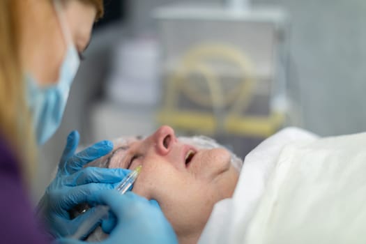 Close-up on the face of a woman lying on a bed in a beauty salon. A cosmetologist is standing next to the bed, inserting a product under the client's skin.
