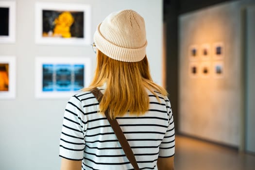 Asian woman standing she looking art gallery in front of colorful framed paintings pictures on white wall, young female watch at photo frame to leaning against at show exhibition gallery, Back view