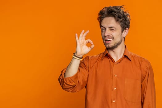 Okay. Happy Caucasian man looking approvingly at camera showing ok gesture like sign positive feedback, approve something good celebrate victory wink blink eye flirting. Young guy on orange background