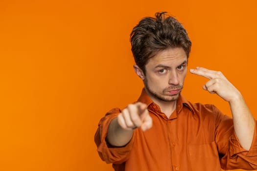 I am watching you. Confident attentive man pointing at his eyes and camera show I am watching you gesture spying on someone. Disappointed Caucasian guy isolated on orange studio background. Copy-space