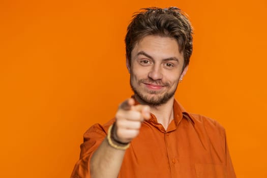 I choose you. Caucasian smiling man pointing finger to camera and looking with playful happy expression making winner choice showing direction. Handsome young guy isolated on orange studio background