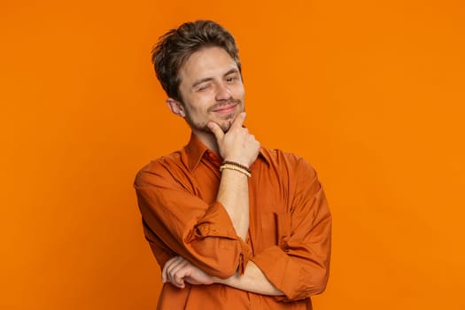 Smiling playful happy Caucasian man blinking eye, looking at camera with smile, winking, flirting, expressing optimism, good news. Charming young adult guy isolated on studio orange background indoors