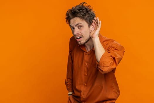 I cant hear you. What. Caucasian man trying hear you looking confused and frowning keeping arm near ear for louder voice asking to repeat, to hear information, deafness. Young guy on orange background