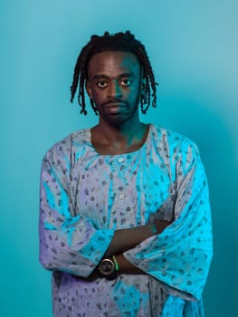 A Sudanese man adorned with modern dreadlocks stands proudly in traditional Sudanese attire, his arms crossed, conveying a blend of cultural heritage and contemporary style against a vibrant blue backdrop