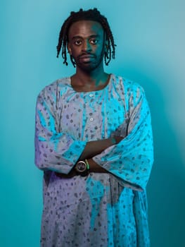 A Sudanese man adorned with modern dreadlocks stands proudly in traditional Sudanese attire, his arms crossed, conveying a blend of cultural heritage and contemporary style against a vibrant blue backdrop