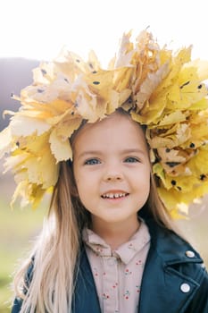 Smiling little girl in a wreath of autumn leaves stands in a meadow. Portrait. High quality photo