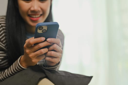 Smiling young asian woman relaxing while chatting, typing message on smartphone.