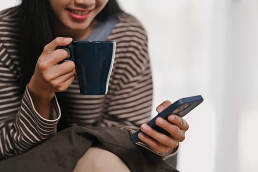 Pleased young woman drinking hot tea and chatting on mobile phone. People and technology concept.