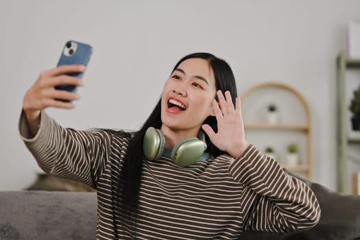 Happy smiling young woman making a selfie or video call to relatives on a sofa in a living room.