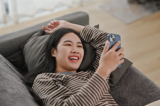 Relaxed young woman chatting or shopping online on mobile phone while lying on couch at home.