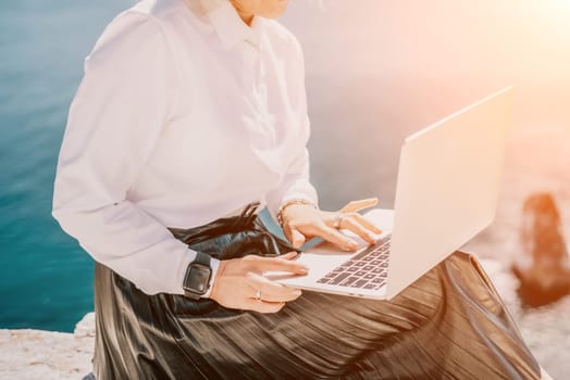 Successful business woman in yellow hat working on laptop by the sea. Pretty lady typing on computer at summer day outdoors. Freelance, travel and holidays concept.