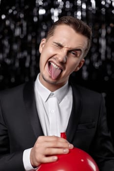 funny guy with red air balloon showing tongue at birthday party on dark background