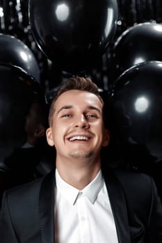 Happy handsome man in black jacket holding black air balloons on glitter background.