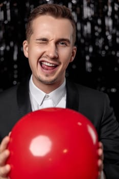 happy man in black jacket with red air balloon winking looking at the camera on dark glitter background.
