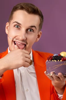 handsome birthday man in orange jacket eating cream cake on purple background
