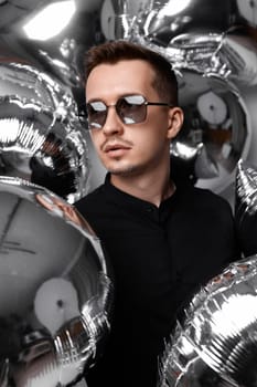 portrait of happy man in black shirt and sunglasses with a lot of silver air balloons background. birthday party