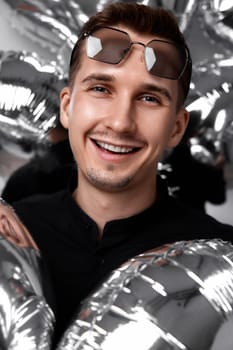 smiling man in black shirt and sunglasses with a lot of silver air balloons background. birthday party