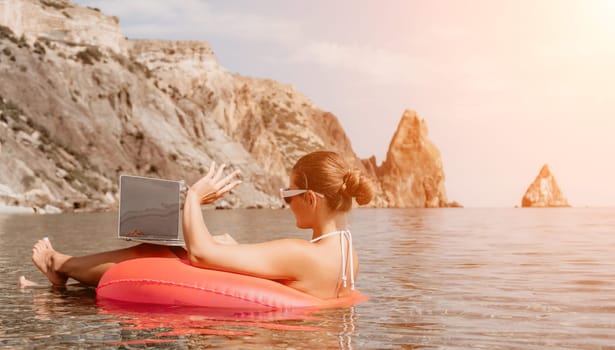 Woman freelancer works on laptop swimming in sea on pink inflatable ring. Pretty lady typing on computer while floating in the sea on inflatable donut at sunset. Freelance, remote work on vacation