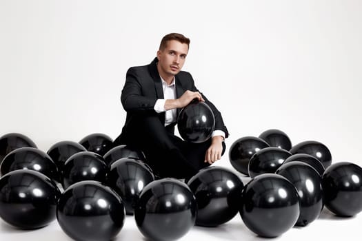 elegant caucasian man in suit tuxedo sitting on studio floor with a lot of black air balloons