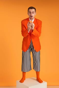 extravagant funny young man in orange jacket and striped pants holding cupcake on orange background.