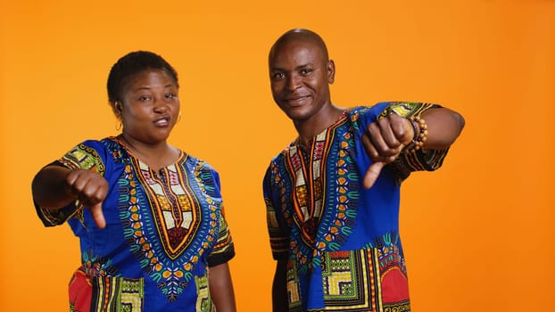 Ethnic man and woman showing thumbs down symbol over orange background, showcasing negative opinion with dislike sign. African american couple presenting disapproval and bad feedback.