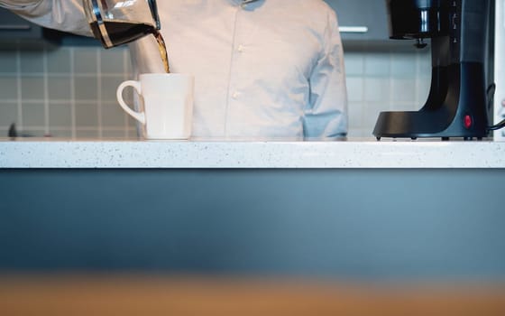 Man in the kitchen pouring a mug of hot filtered coffee from a glass pot in the morning