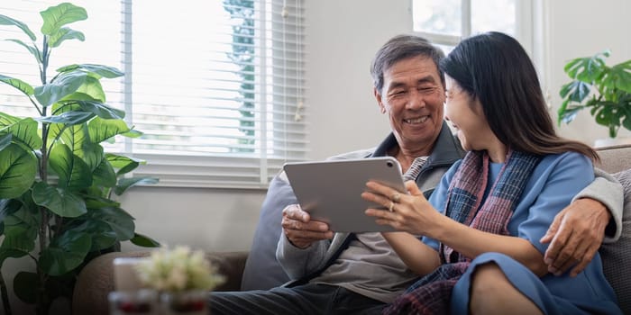 Happy elderly asian couple using tablet sit on sofa doing ecommerce shopping online on website and buying insurance browsing at home.