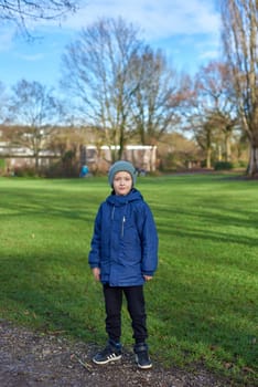 Young Autumn Explorer: Beautiful 8-Year-Old Boy in Jacket and Beanie Standing in the Park with Green Lawn. Embark on a journey of youthful wonder with this captivating image featuring a beautiful 8-year-old boy, clad in a jacket and beanie, standing in the autumn park. The lush green lawn provides a vibrant canvas for the charm of fall, creating a perfect blend of innocence and seasonal beauty.