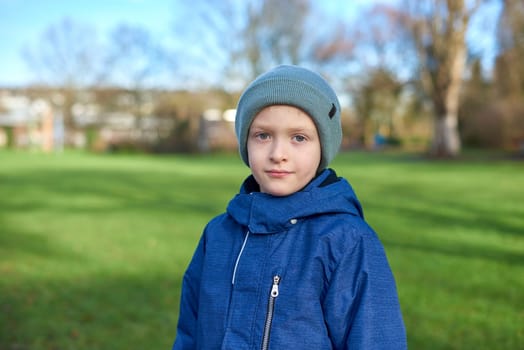 Young Autumn Explorer: Beautiful 8-Year-Old Boy in Jacket and Beanie Standing in the Park with Green Lawn. Embark on a journey of youthful wonder with this captivating image featuring a beautiful 8-year-old boy, clad in a jacket and beanie, standing in the autumn park. The lush green lawn provides a vibrant canvas for the charm of fall, creating a perfect blend of innocence and seasonal beauty.