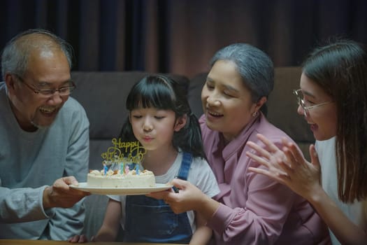 Happy Asian family grandparents and mother celebrating enjoy birthday party with cake of granddaughter Blowing candle at home in night time, Birthday surprise concept