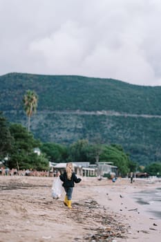 Little girl with a bag walks along a dirty beach collecting trash. Back view. High quality photo