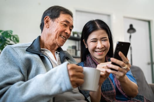 Retired elderly couple sits on couch drink tea and using mobile together and relax in their home. Senior Activity Concept.