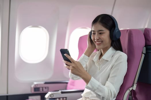 Cheerful Asian woman passenger in headphone watching online movie during intercontinental flight in cabin of aircraft, happy traveler using wifi connection on board.