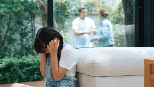 Stressed and unhappy young girl huddle in corner, cover her ears blocking sound of her parent arguing in background. Domestic violence at home and traumatic childhood develop to depression. Synchronos